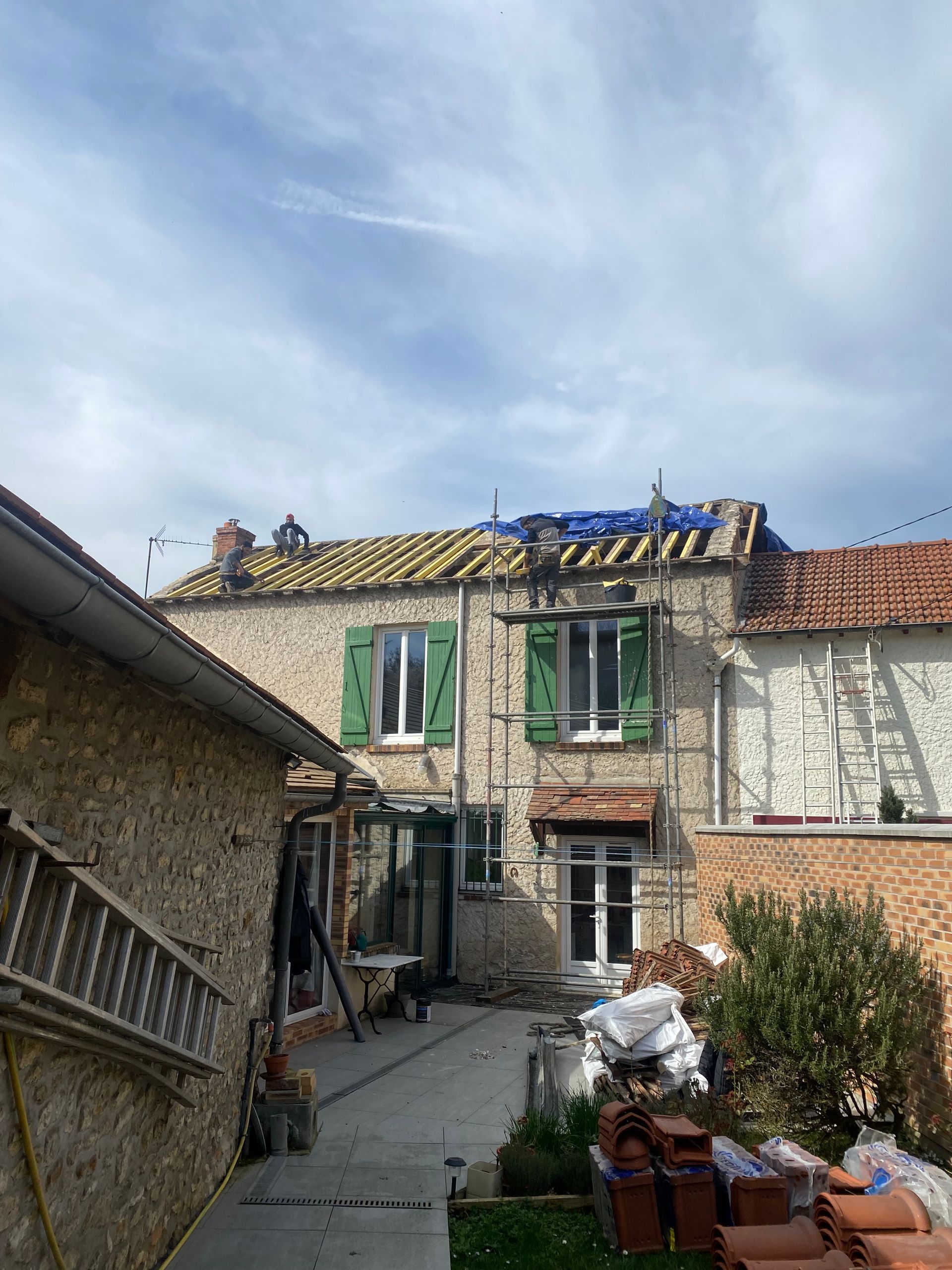 Maison en construction avec tuiles manquantes, volets verts et échafaudages.