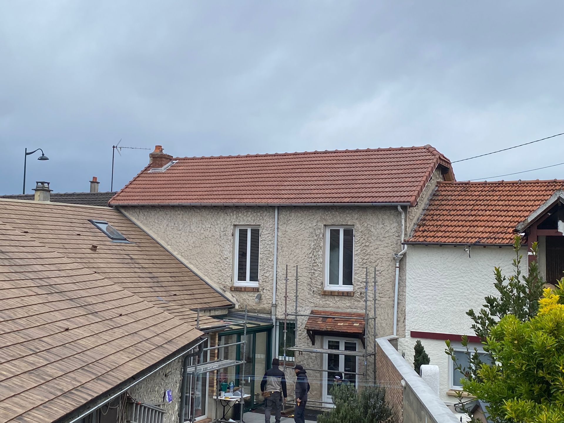 Maison à deux étages avec toit de tuiles rouges et murs en stuc beige. Deux personnes se tiennent près d'une entrée couverte. Ciel couvert.