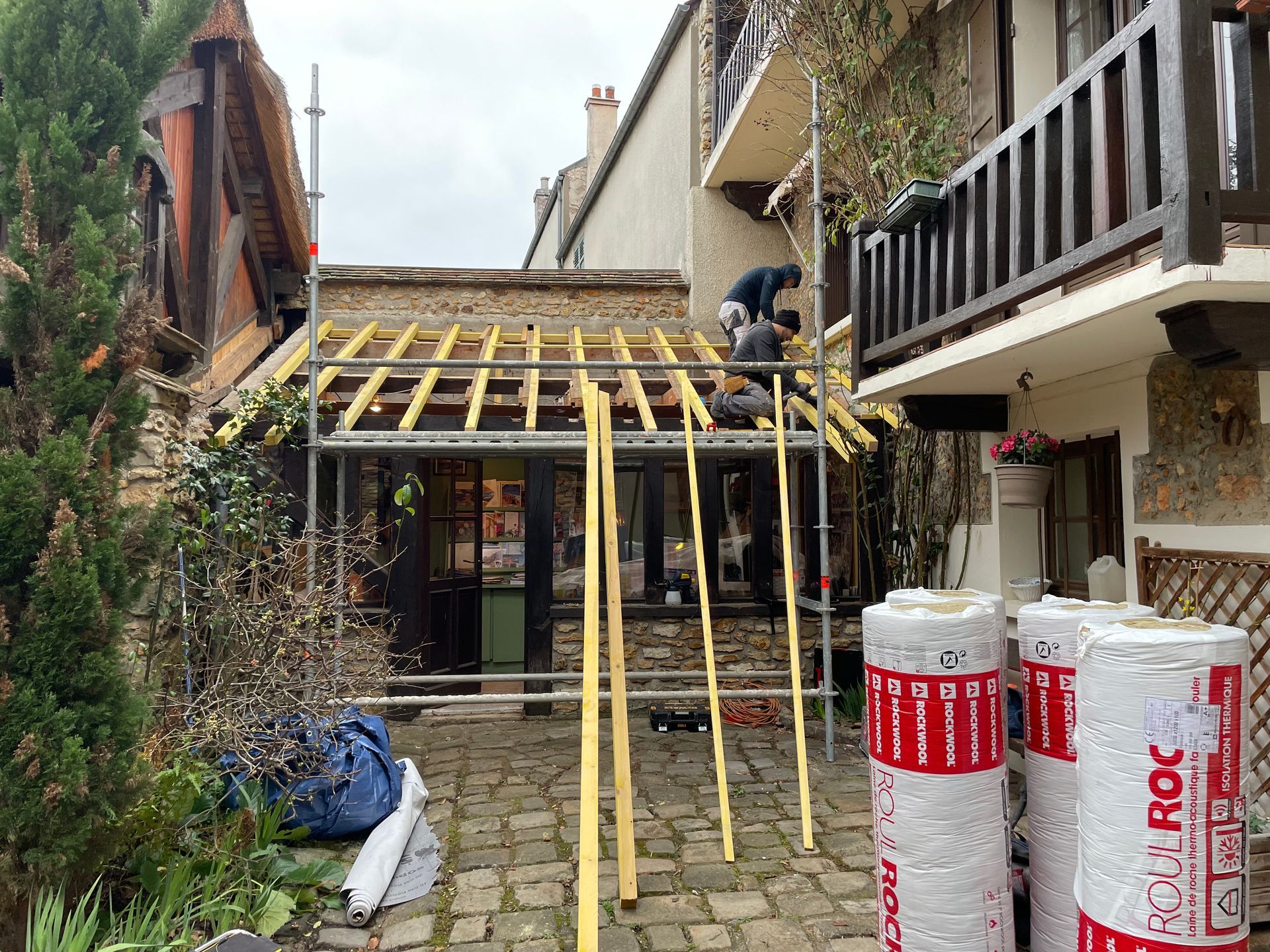 Des ouvriers du bâtiment sur un toit construisent une structure au-dessus d'une porte. Des rouleaux d'isolant sont au premier plan.