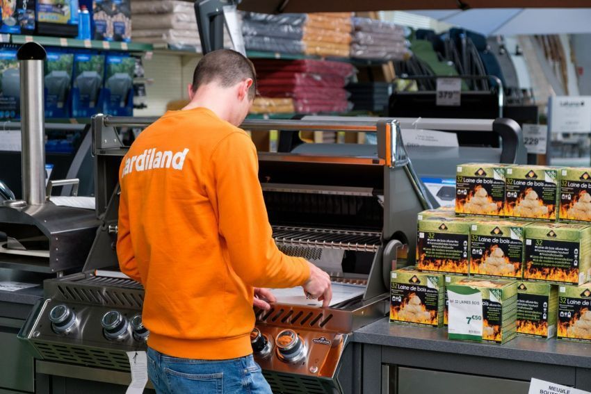 Une personne fait des grillades devant un présentoir de barbecues dans un supermarché, avec des aliments emballés et des accessoires empilés.