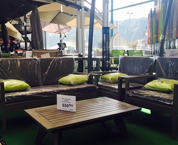 Salon de jardin avec canapés en osier foncé, coussins verts et table en bois sous parasols.