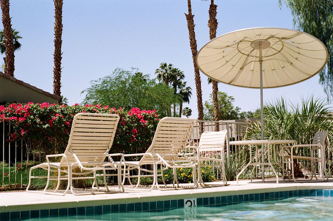 Chaises longues au bord de la piscine sous un grand parasol blanc, à côté de fleurs roses et de palmiers