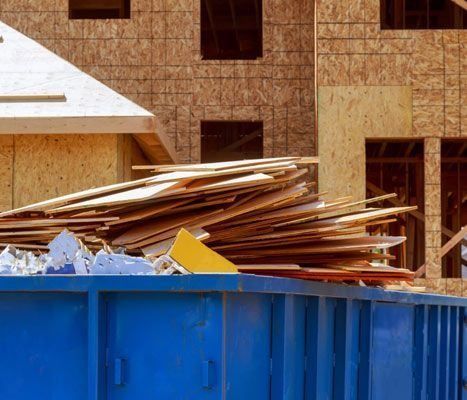 Un contenedor de basura azul lleno de madera está frente a una casa en construcción.