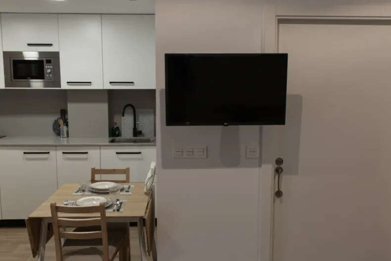Cocina con armarios blancos, microondas, televisor montado en la pared y mesa de comedor puesta para comer.