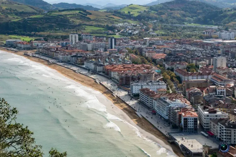 Paisaje urbano costero con playa, edificios y montañas al fondo bajo un cielo azul.