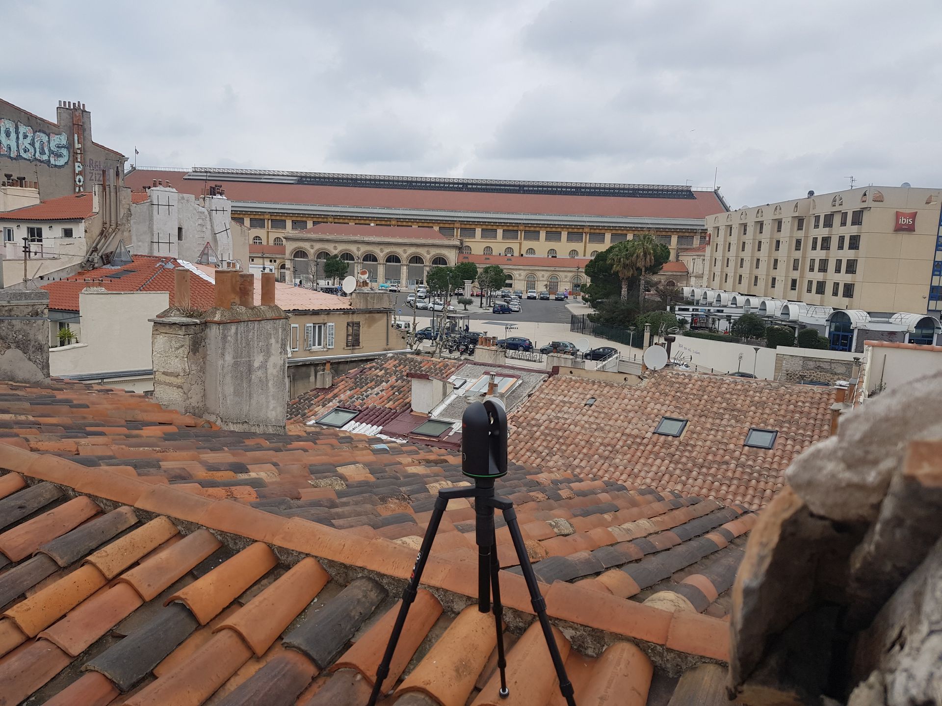 Caméra sur trépied au sommet d'un toit de tuiles rouges, surplombant un paysage urbain avec des bâtiments et des véhicules stationnés.