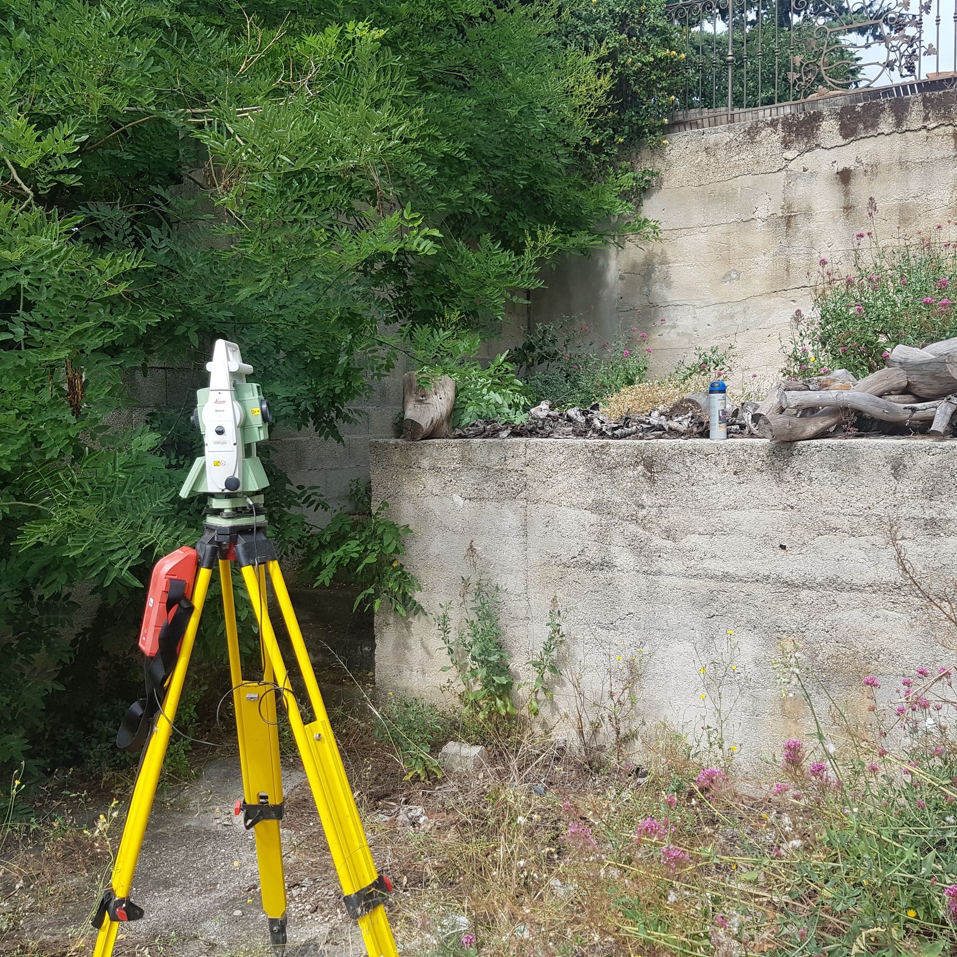Un instrument de topographie sur un trépied à côté d'un mur en béton et d'arbres.