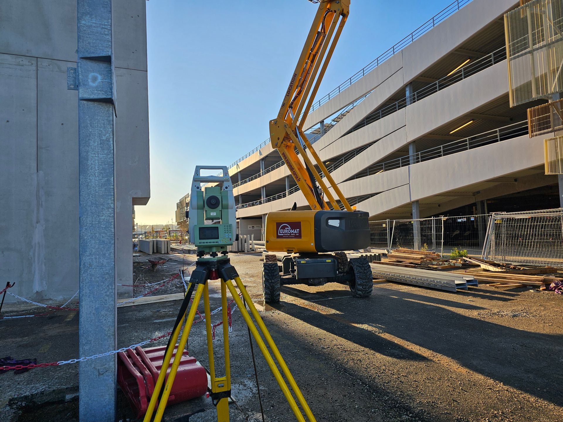 Un géomètre, portant un casque et un gilet de sécurité, utilise un théodolite sur un chantier. Un autre ouvrier se tient à l'arrière-plan.