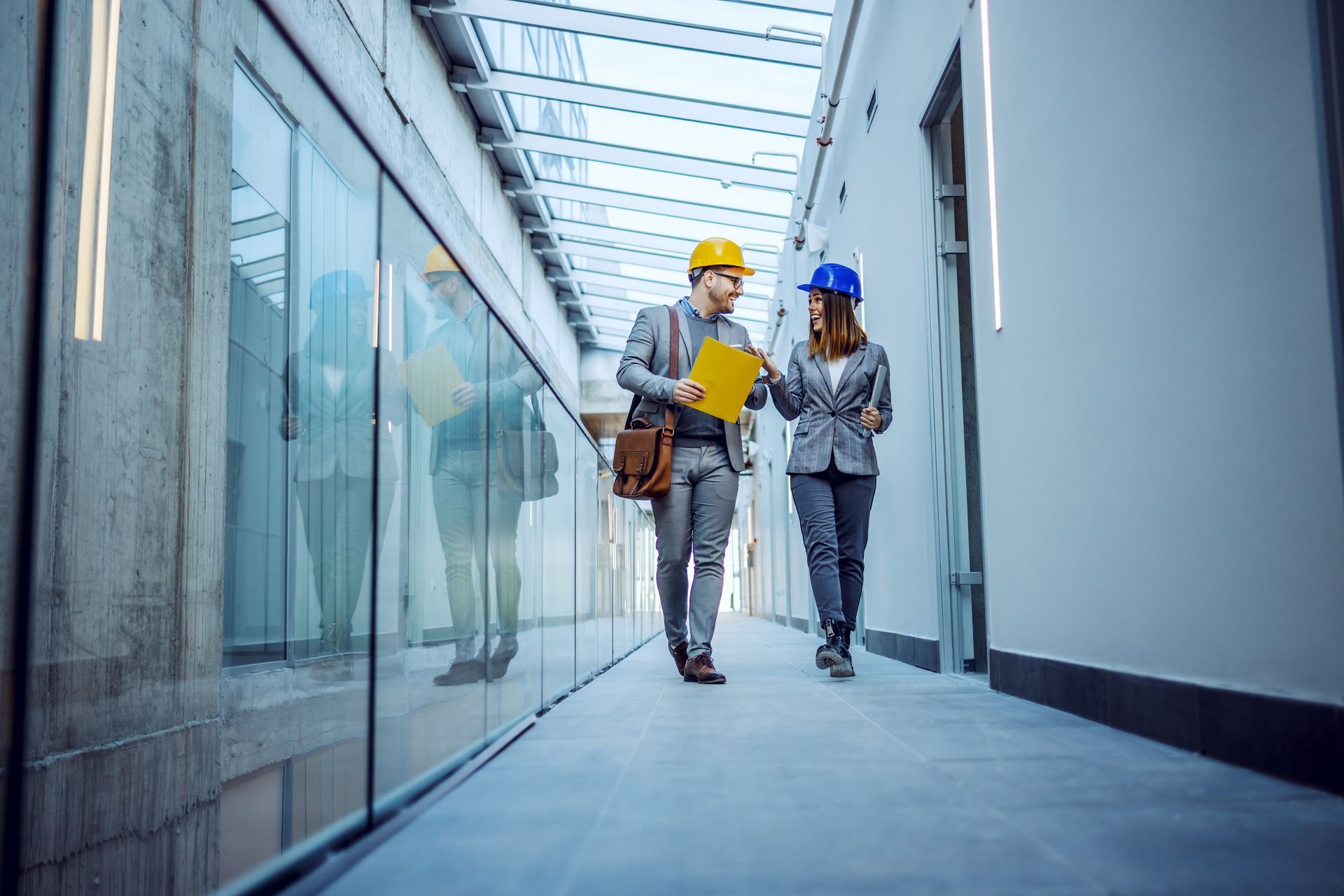 Deux personnes portant des casques de chantier marchent et discutent de documents dans un couloir moderne aux parois vitrées et doté d'une verrière.