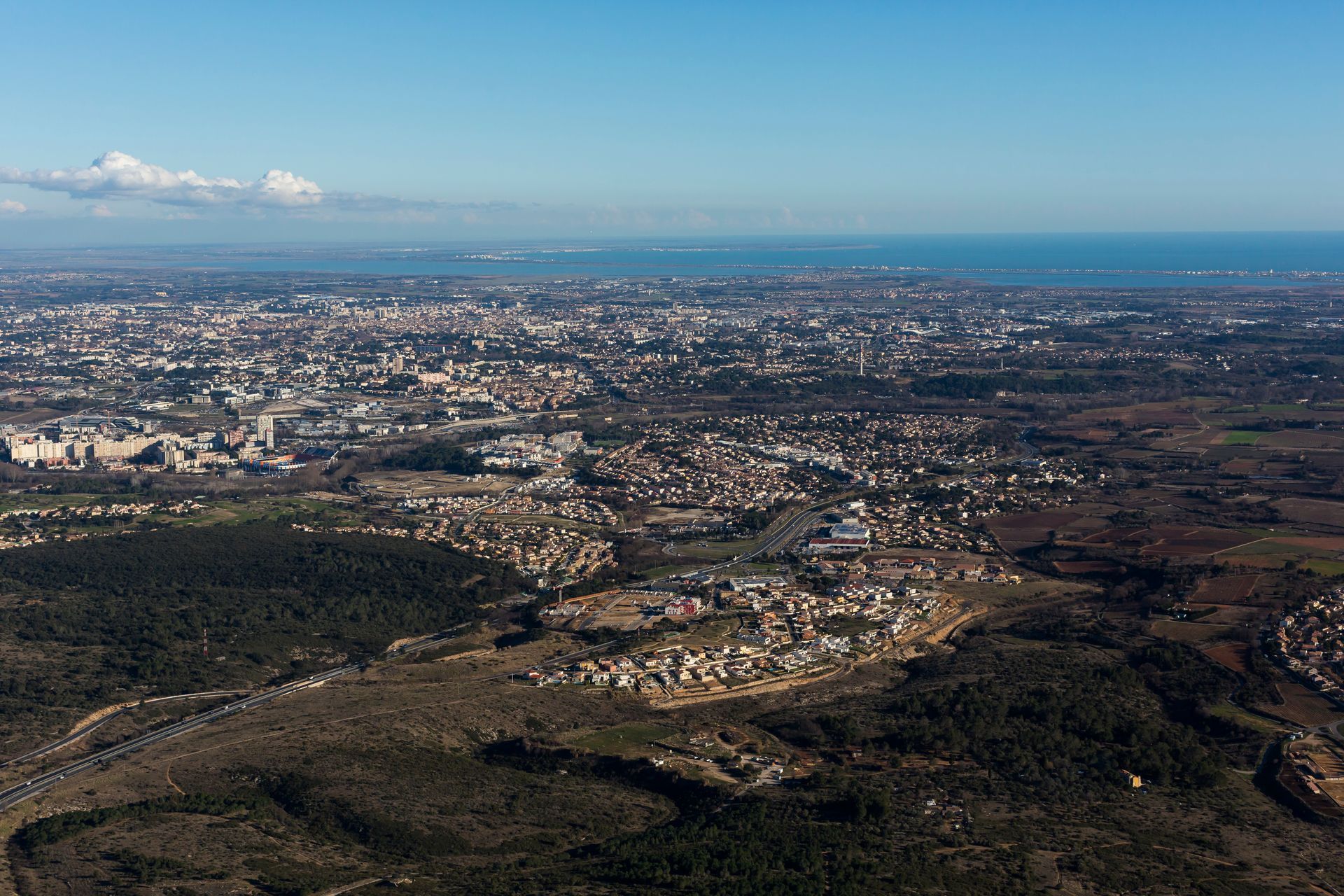 Vue aérienne d'une ville par une journée ensoleillée, avec des bâtiments disséminés dans le paysage et une étendue d'eau au loin.