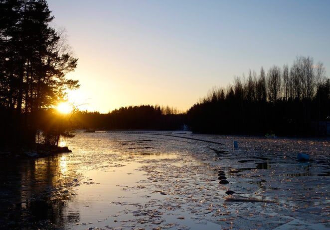 Auringonlasku osittain jäätyneen järven yllä, puiden siluetit rannalla