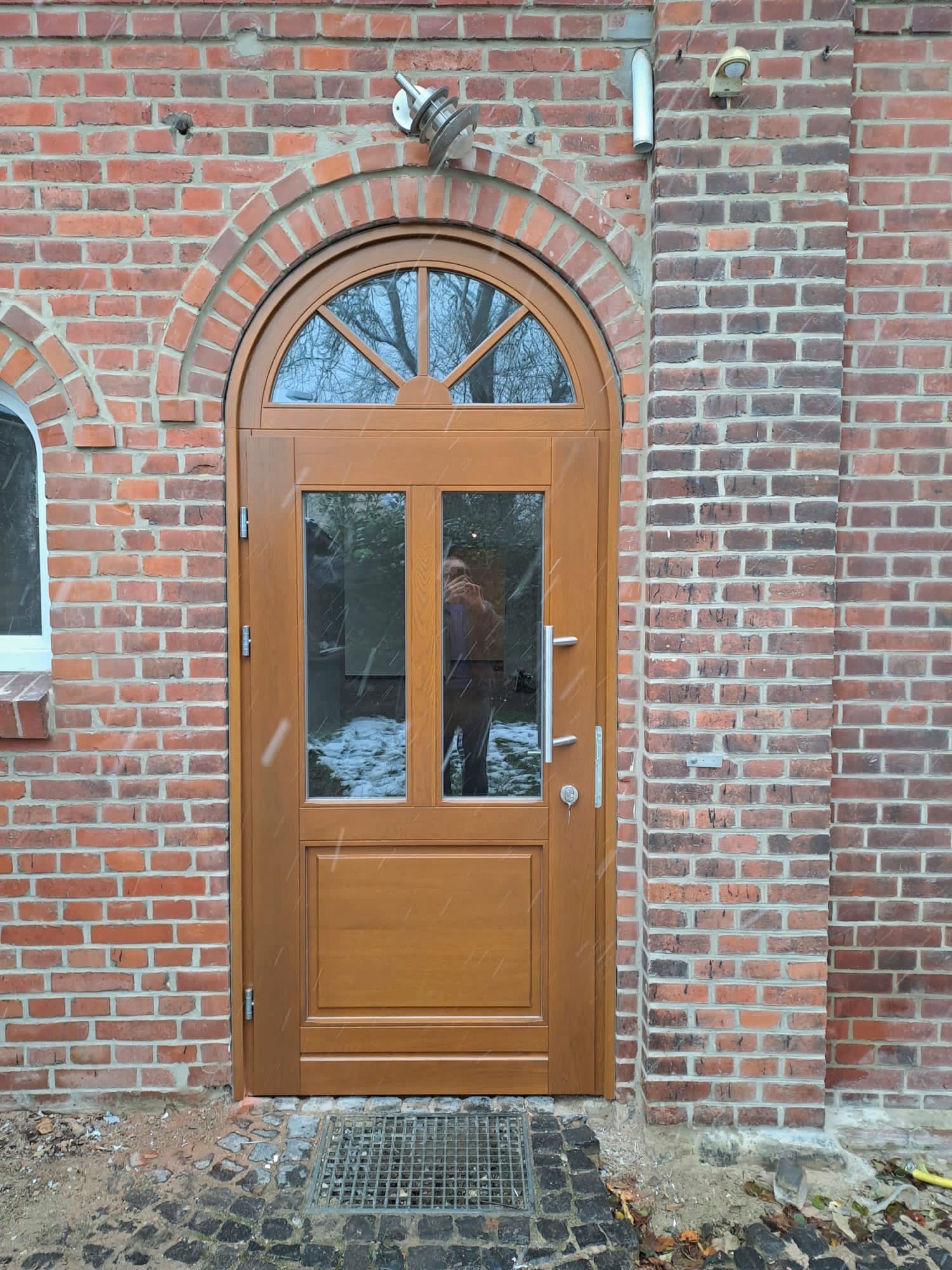 An einem Backsteingebäude befindet sich eine Holztür mit rundem Fenster.