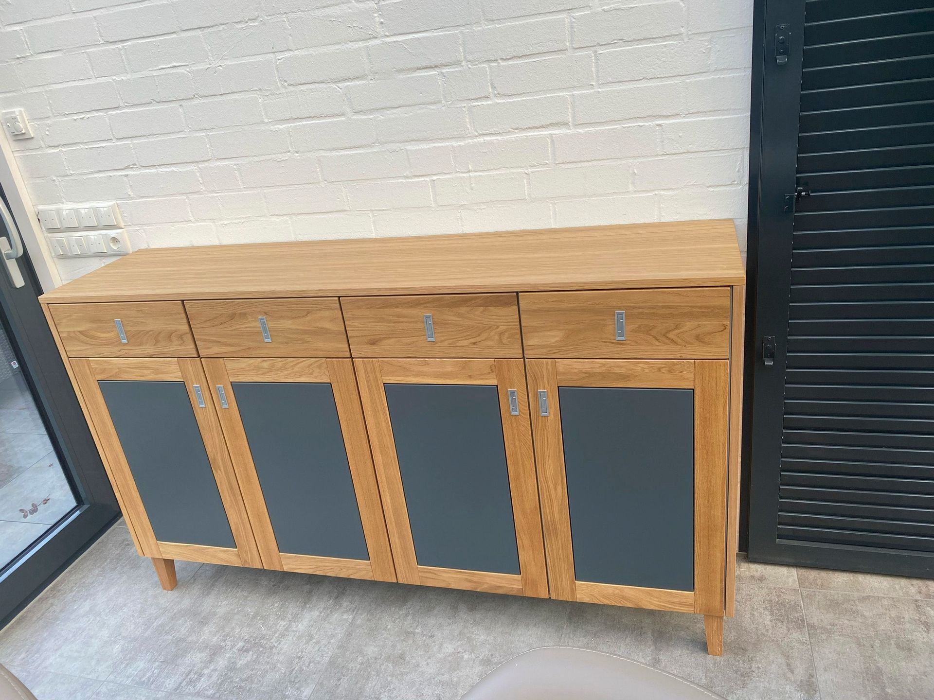 Vor einer weißen Ziegelwand steht ein Holzschrank mit grauen Türen und Schubladen.