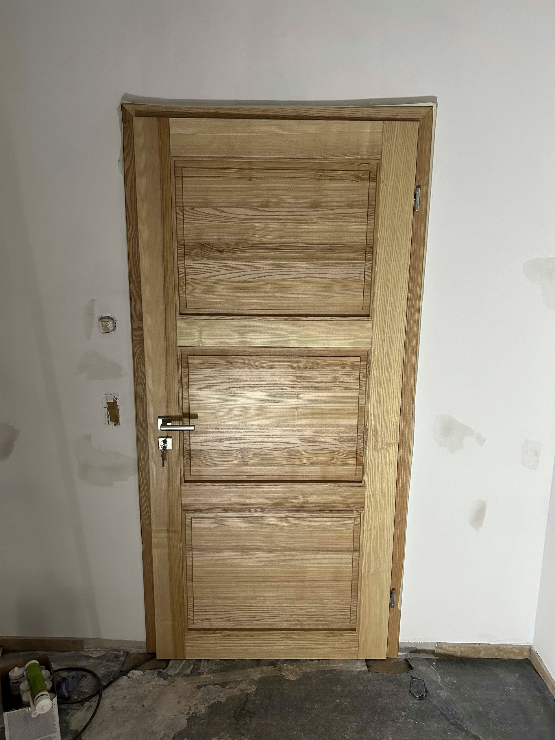 In einem Raum neben einer weißen Wand befindet sich eine Holztür.