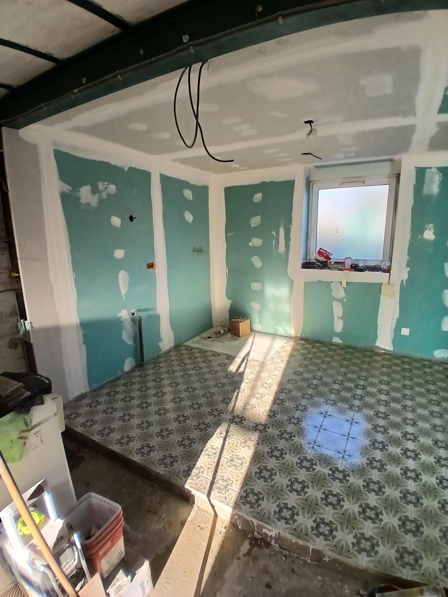 Pièce vide en rénovation avec cloisons sèches vertes, sol à motifs, câbles apparents et lumière naturelle grâce à une fenêtre.