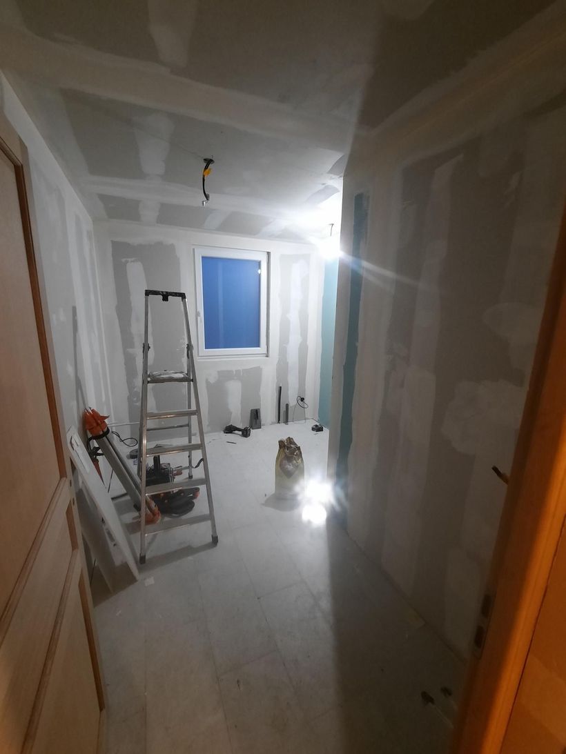 Couloir étroit en cours de rénovation, avec cloisons sèches, échelles, outils et une fenêtre au fond.