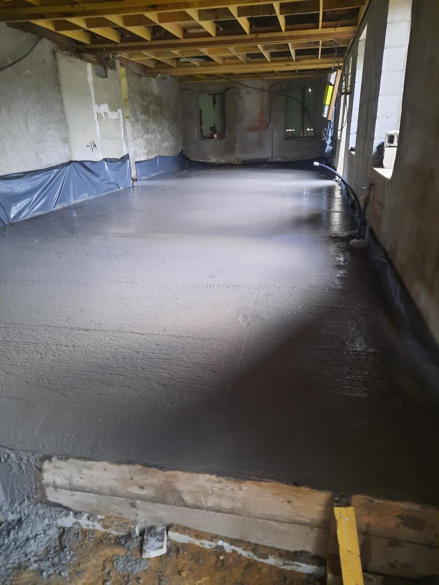 Sol en béton fraîchement coulé dans une pièce intérieure inachevée, avec poutres apparentes et murs recouverts de plastique.
