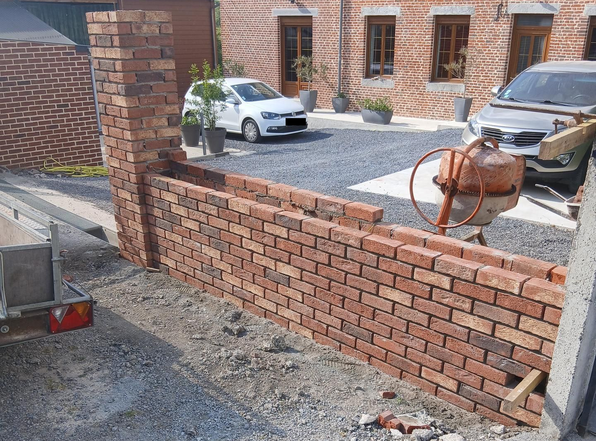 Construction en cours d'un muret en brique avec des voitures, une bétonnière et une maison en brique en fond.