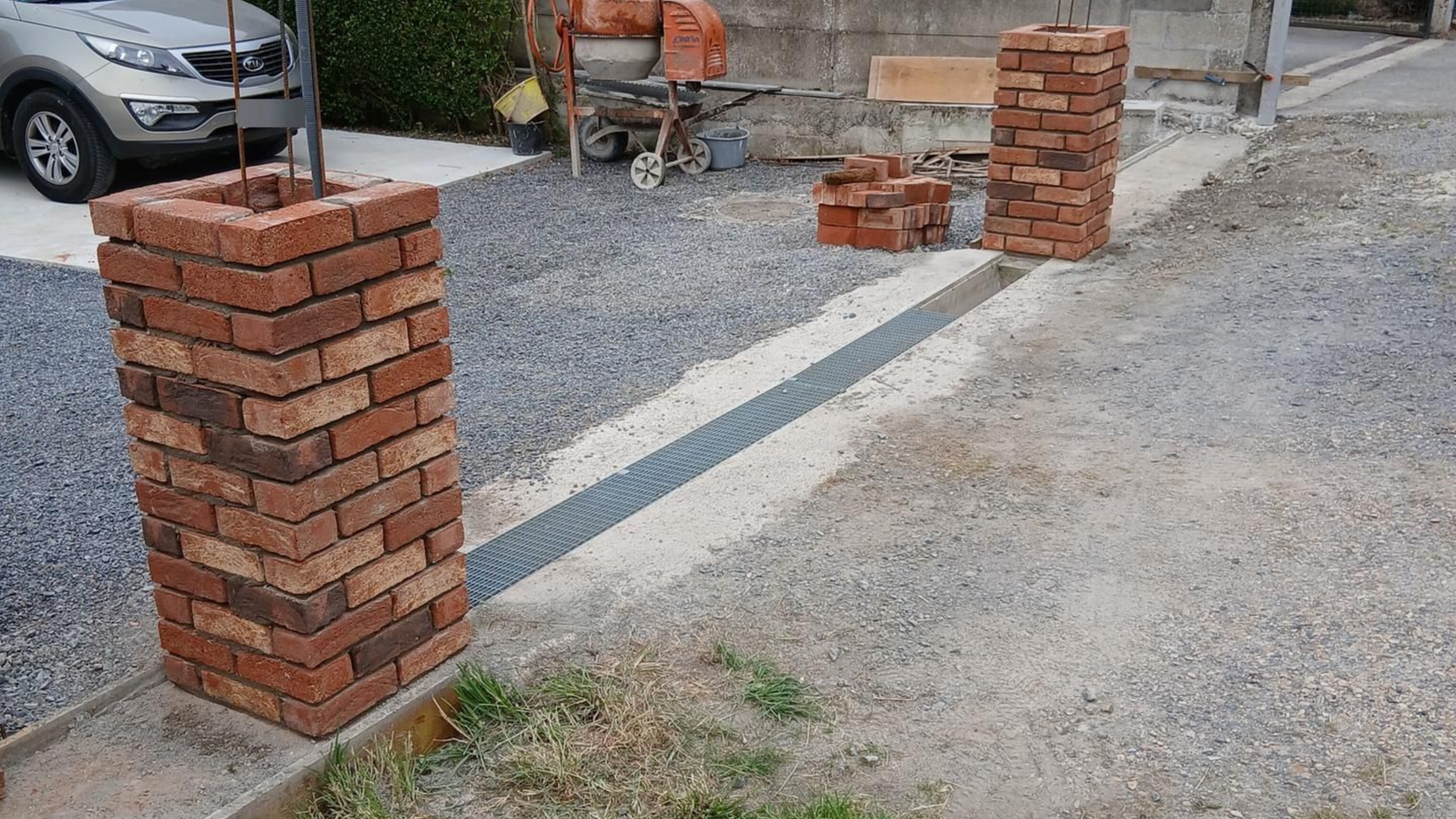 Des piliers en briques sont en construction le long d'un trottoir, à côté d'une allée de gravier, avec des briques empilées et une voiture garée.