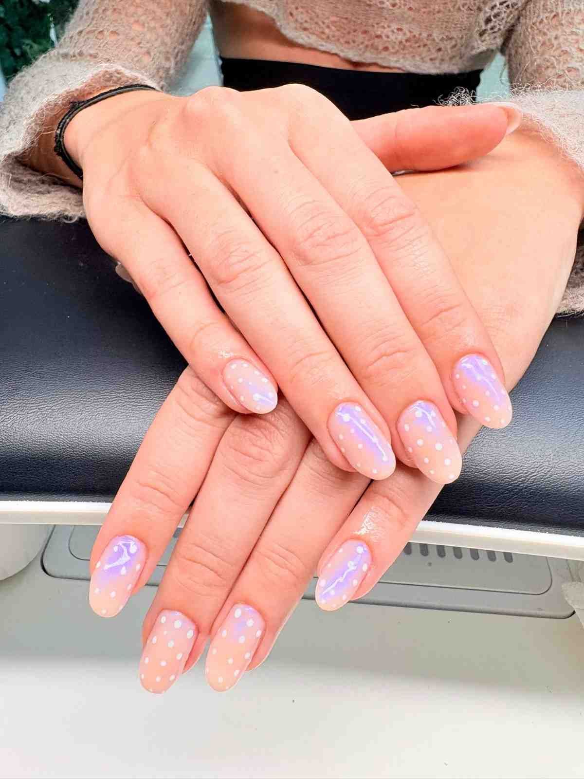 Manos con uñas de forma ovalada, de color rosa pálido con lunares blancos y un toque de brillo morado.