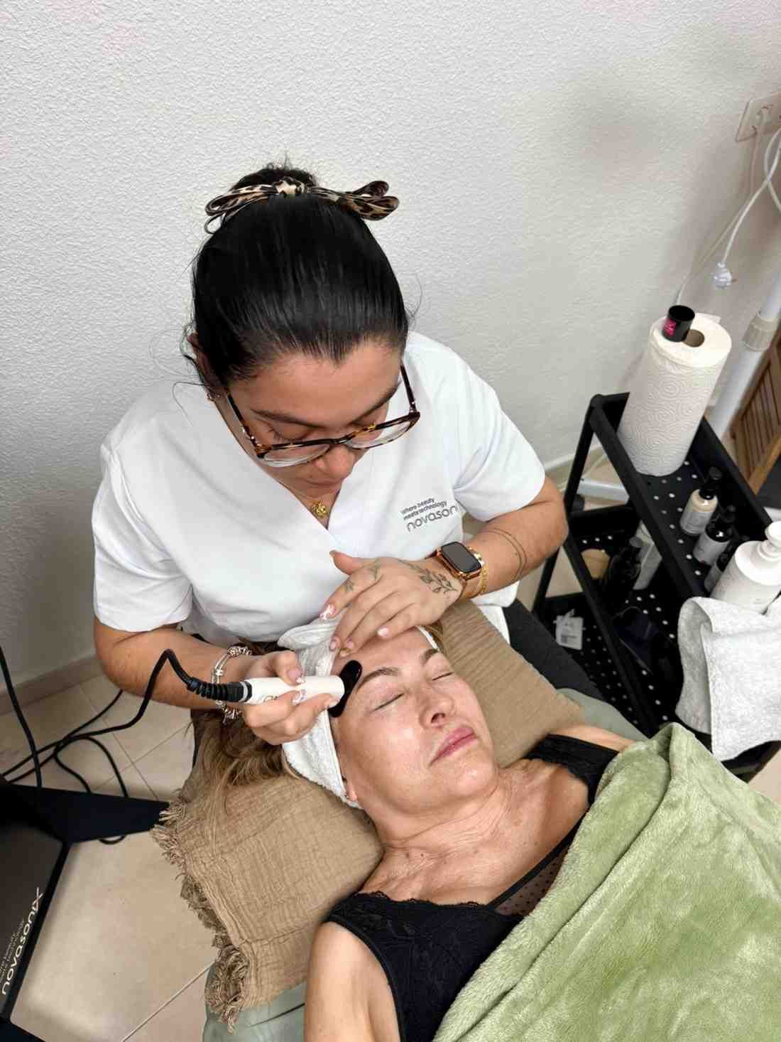 Mujer recibiendo tratamiento facial por parte de una persona con gafas y uniforme blanco. El entorno es un spa.