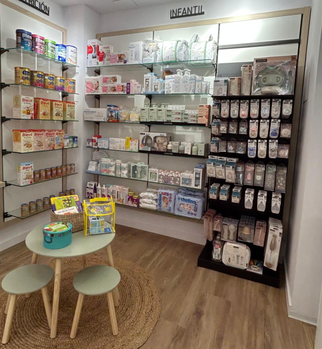 Un farmacéutico con bata blanca sonríe y explica la medicación a un cliente en la farmacia. Estantes de productos al fondo.