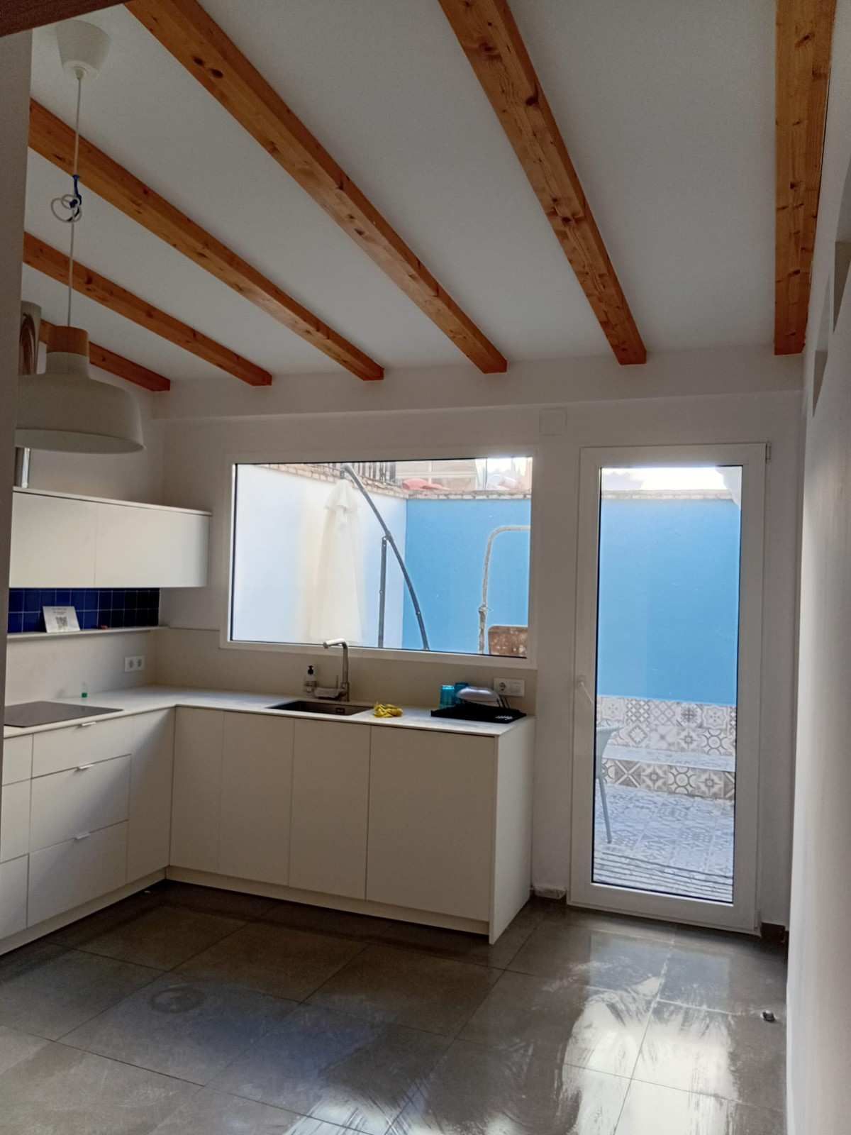 Cocina luminosa con armarios blancos, vigas de madera a la vista en el techo y una puerta de cristal que da a un patio azul.