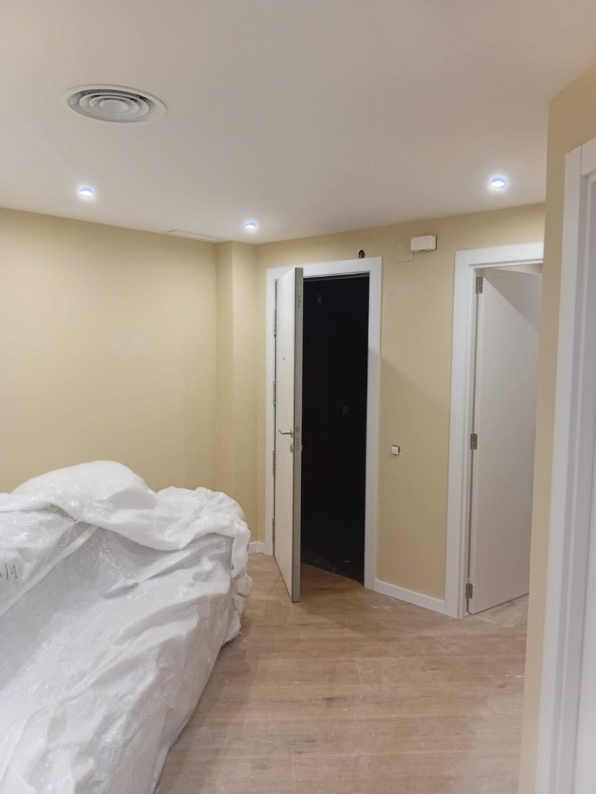 Habitación pequeña de color beige con una cama sin hacer y puertas abiertas que dan a un pasillo oscuro y un baño.