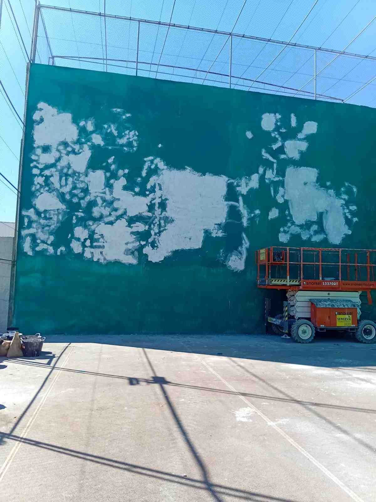 Gran pared turquesa con la pintura desconchada y un pequeño elevador estacionado al lado en un aparcamiento soleado.