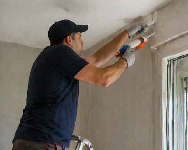 Un pintor aplica pintura blanca en la esquina de una pared con un rodillo, subido a una escalera.
