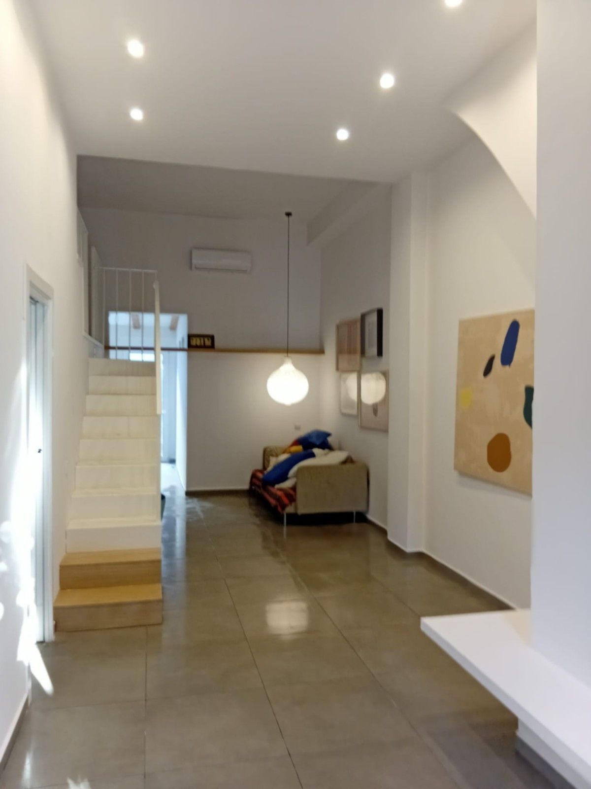 Habitación moderna de color blanco luminoso con escaleras, sofá, cuadros en las paredes y luces empotradas en el techo.