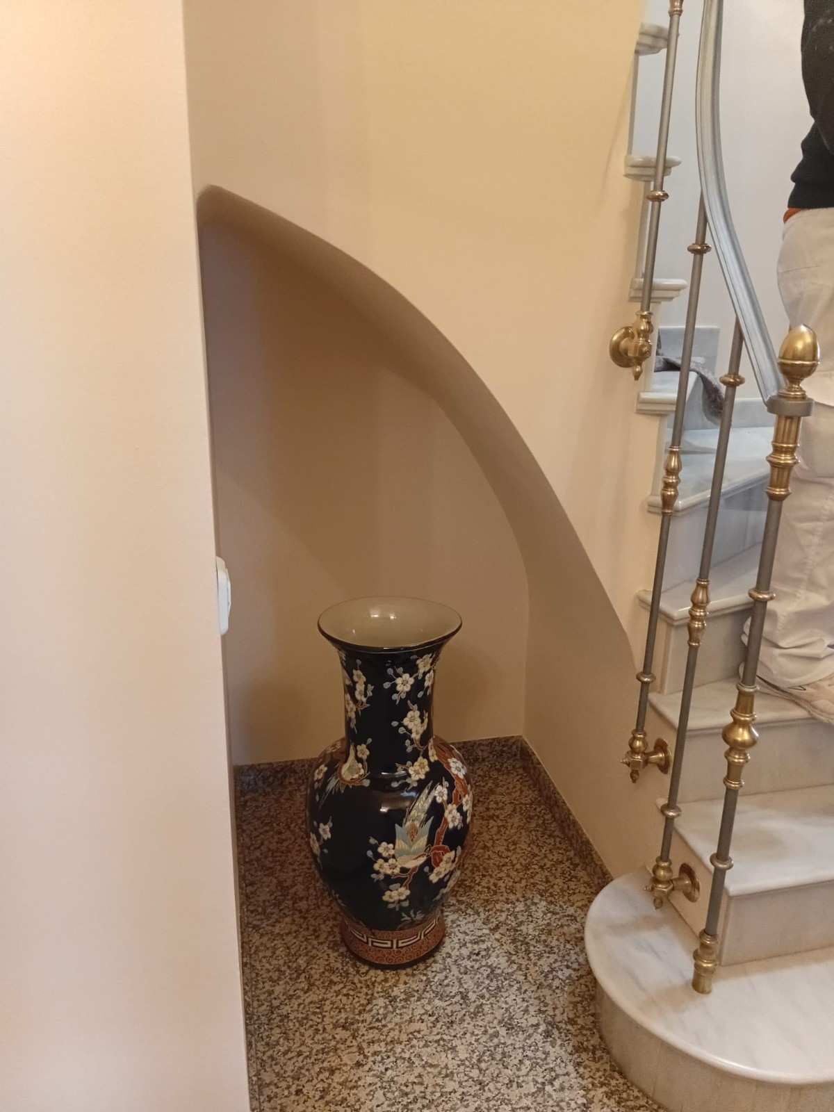 Jarrón decorativo con flores en una pequeña hornacina junto a una escalera con barandilla dorada y blanca.