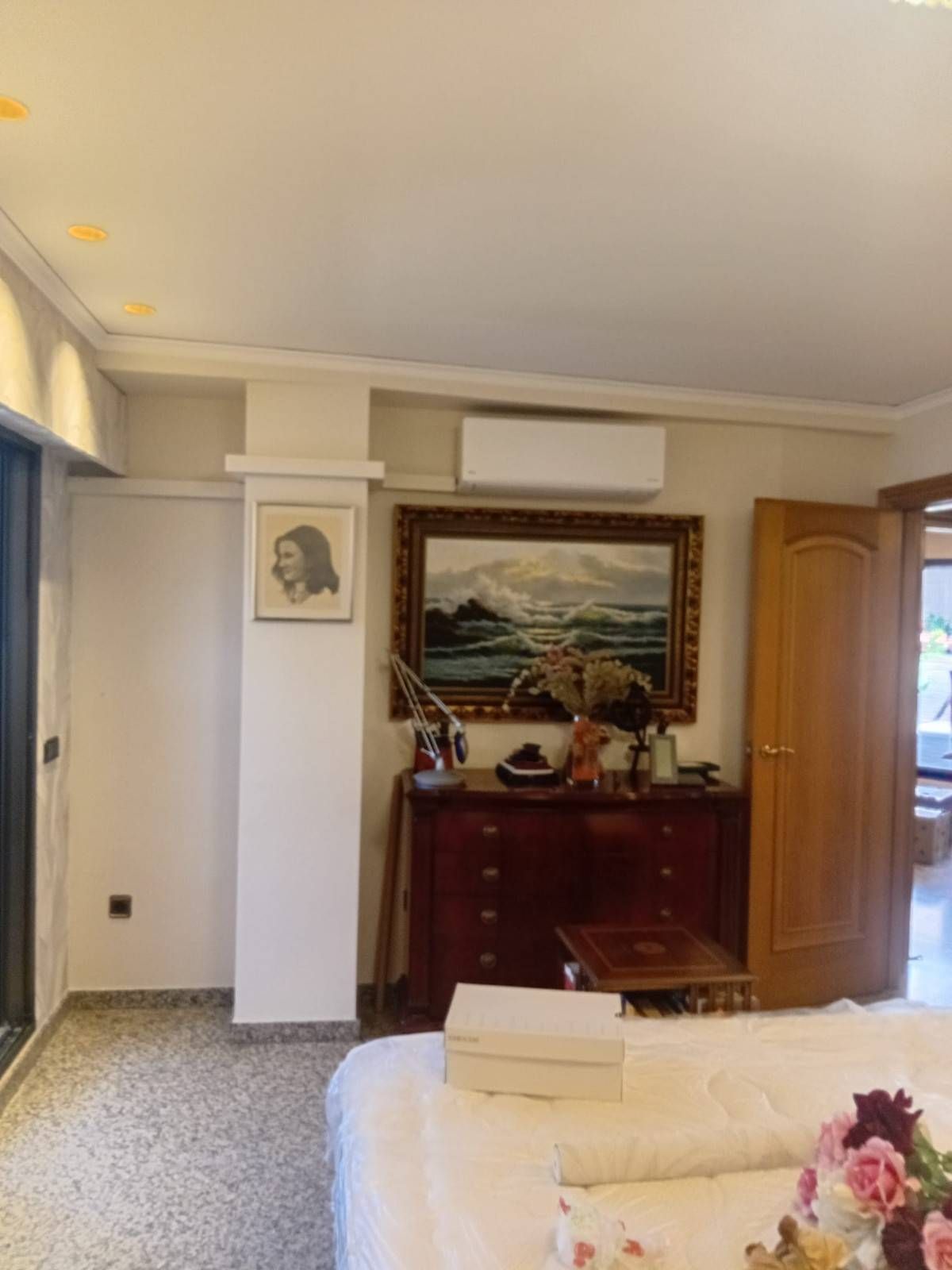 Dormitorio con cama, cómoda, cuadros enmarcados y aire acondicionado cerca de una puerta de madera.