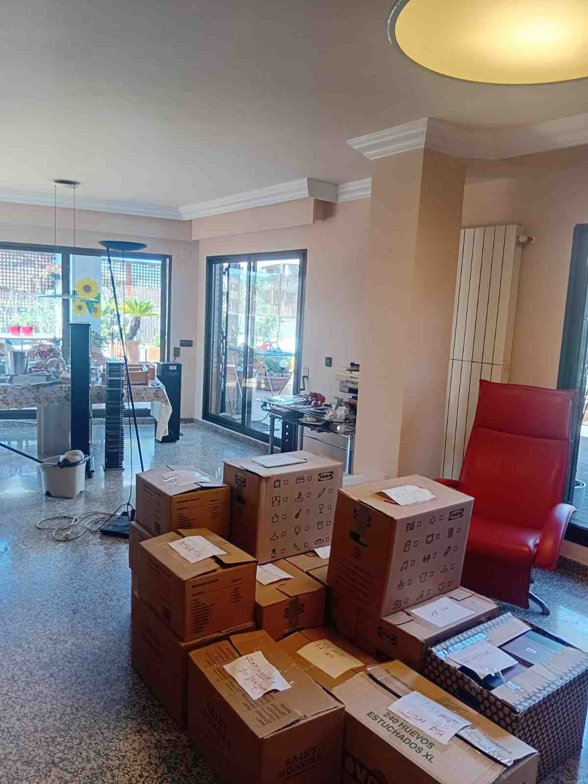Sala de estar con cajas de mudanza apiladas, silla roja y puertas corredizas de vidrio que dan a un patio.
