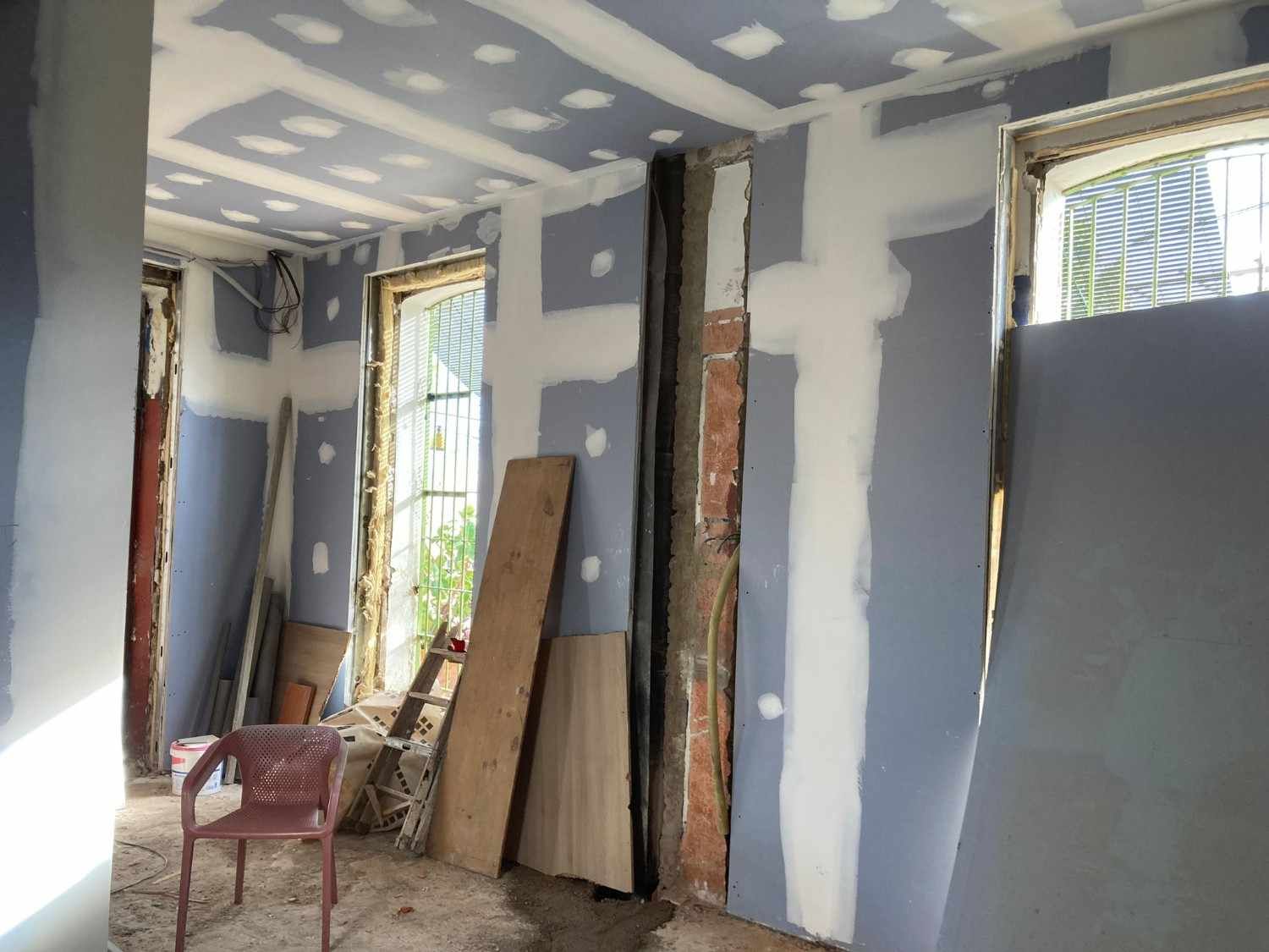 Una persona pinta una pared de blanco en una habitación luminosa con una escalera y materiales de pintura cerca.