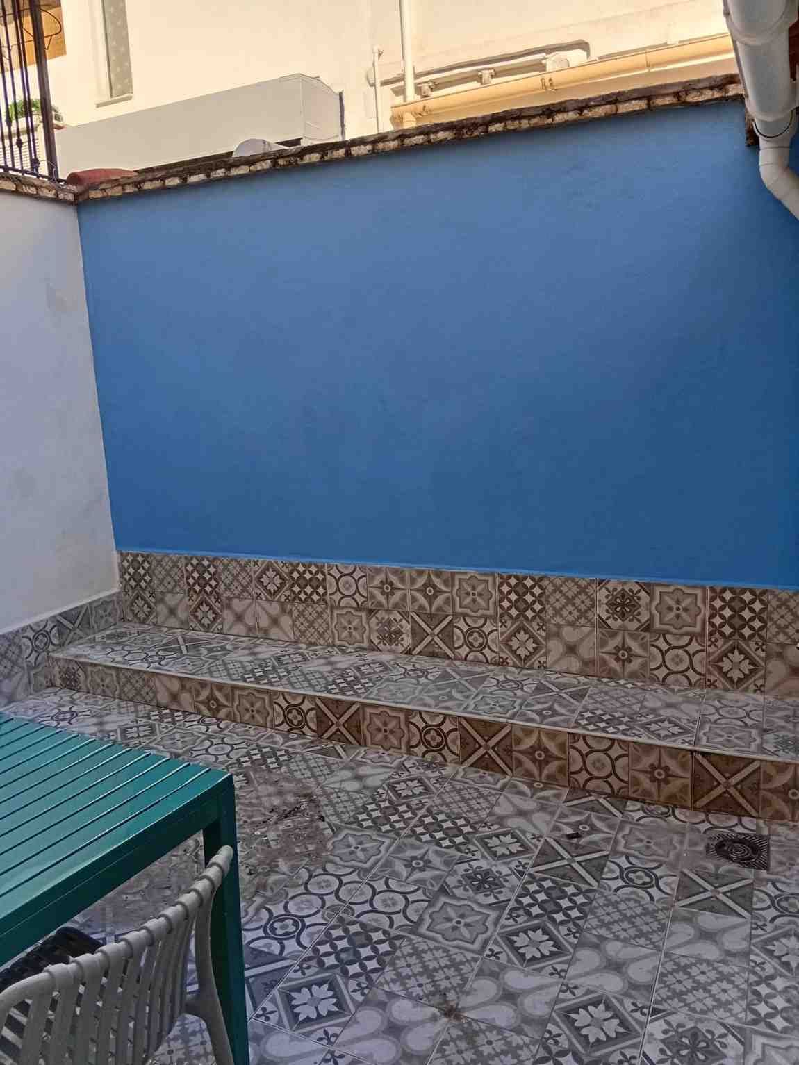 Pared azul con banco y suelo de baldosas estampadas junto a una mesa y una silla verdes en un patio.