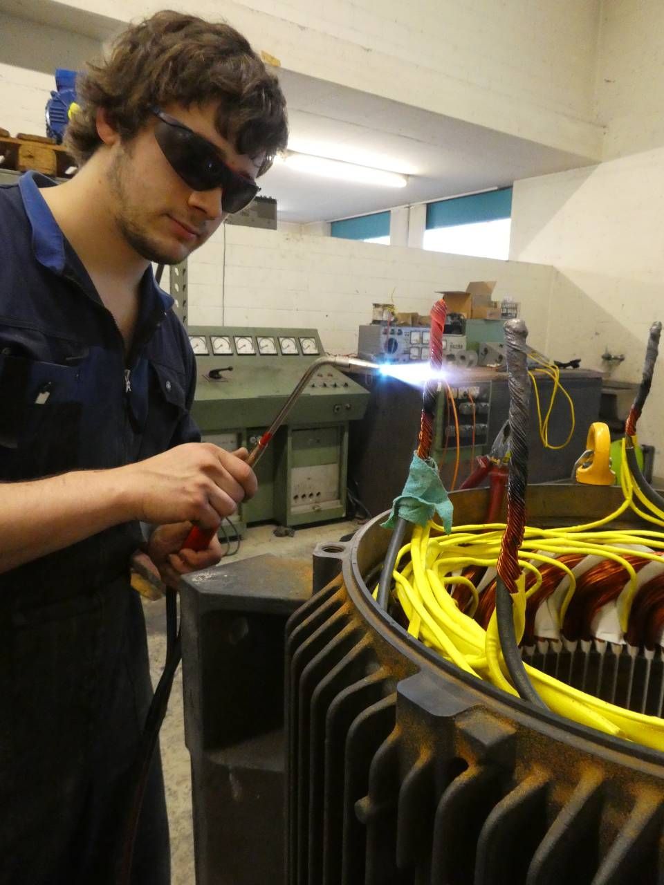 Techniker arbeitet in einer Werkstatt neben einer elektrischen Maschine mit Kabeln und trägt dabei eine Schutzbrille.