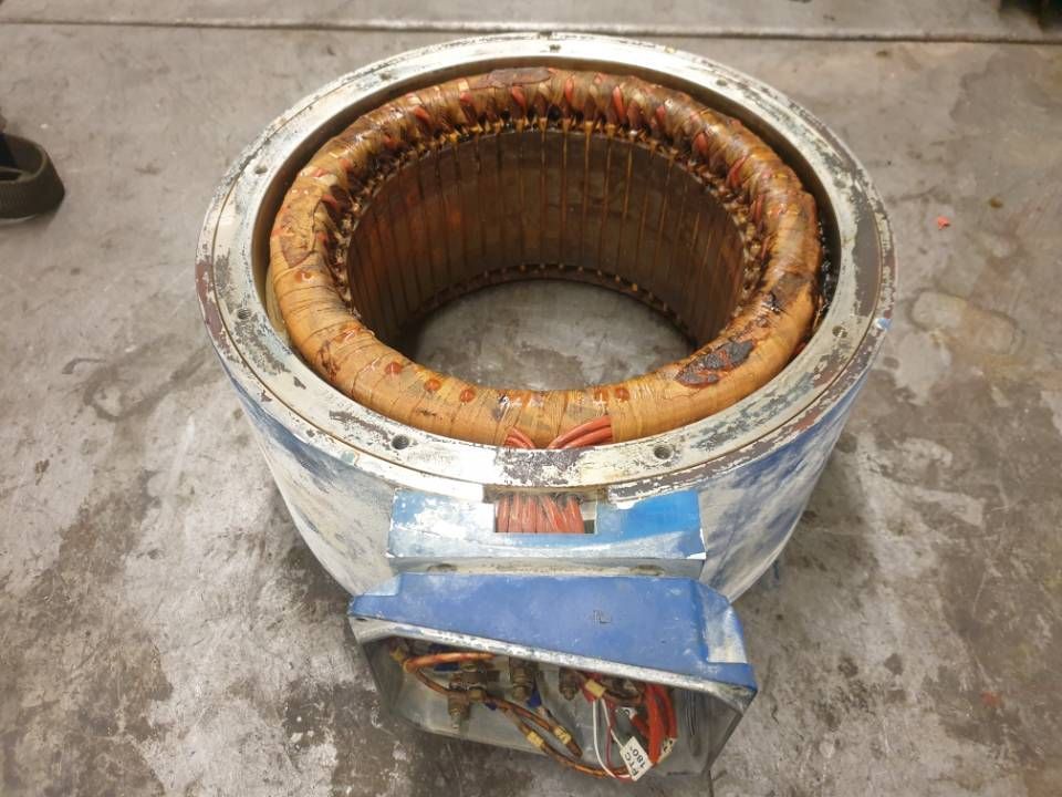 Beschädigte Transformatorspule aus Metall mit freiliegenden Kupferwicklungen auf einem Betonboden