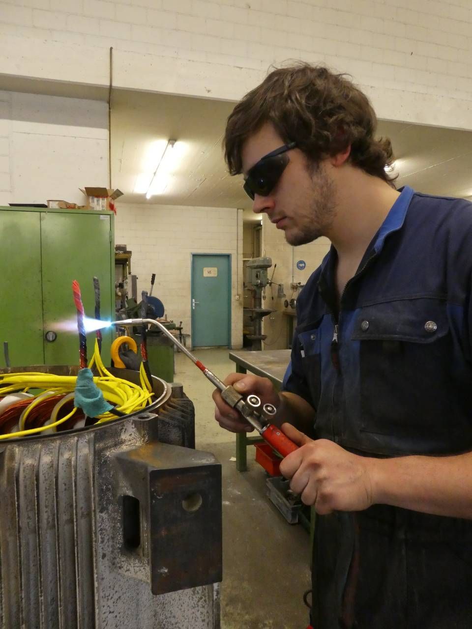 Ein Mann in blauem Overall benutzt eine Zange in der Nähe einer Werkbank in einer Industriewerkstatt.