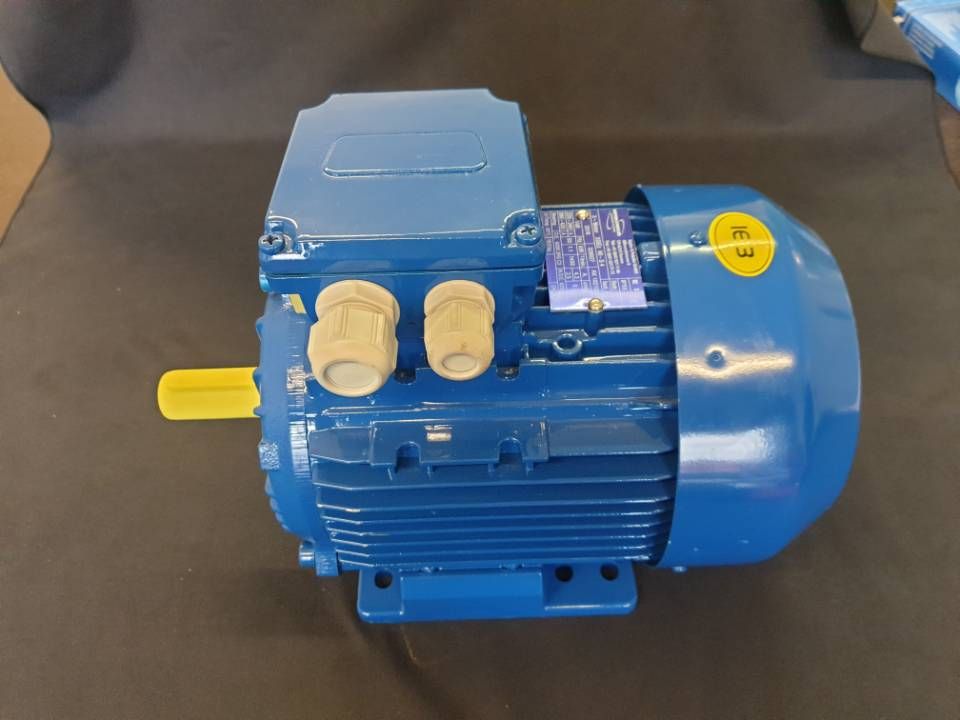 Blauer Elektromotor auf grauer Oberfläche, mit gelber Welle und cremefarbenen Schaltkästen.