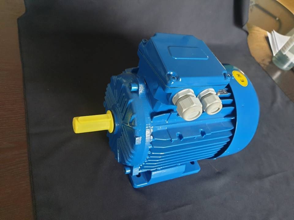 Blauer Elektromotor mit gelber Welle und silbernen Beschlägen auf dunklem Untergrund.