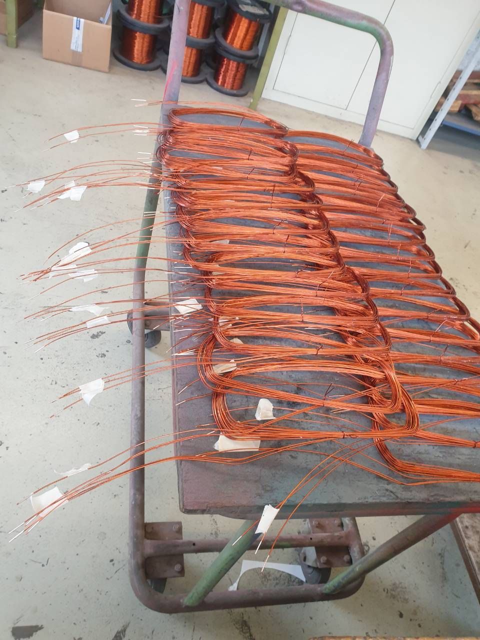 In einer Werkstatt befinden sich orangefarbene Kupferspulen auf einem Metallwagen, von einer Seite ragen Drähte hervor.