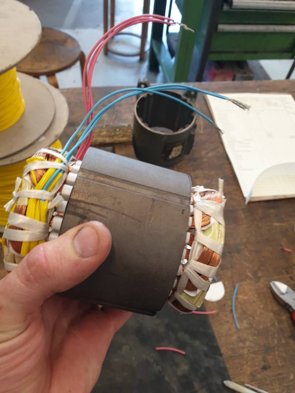 Eine Hand hält eine zylindrische elektrische Spule mit sichtbaren Kupferwicklungen auf einem Werkstatttisch.