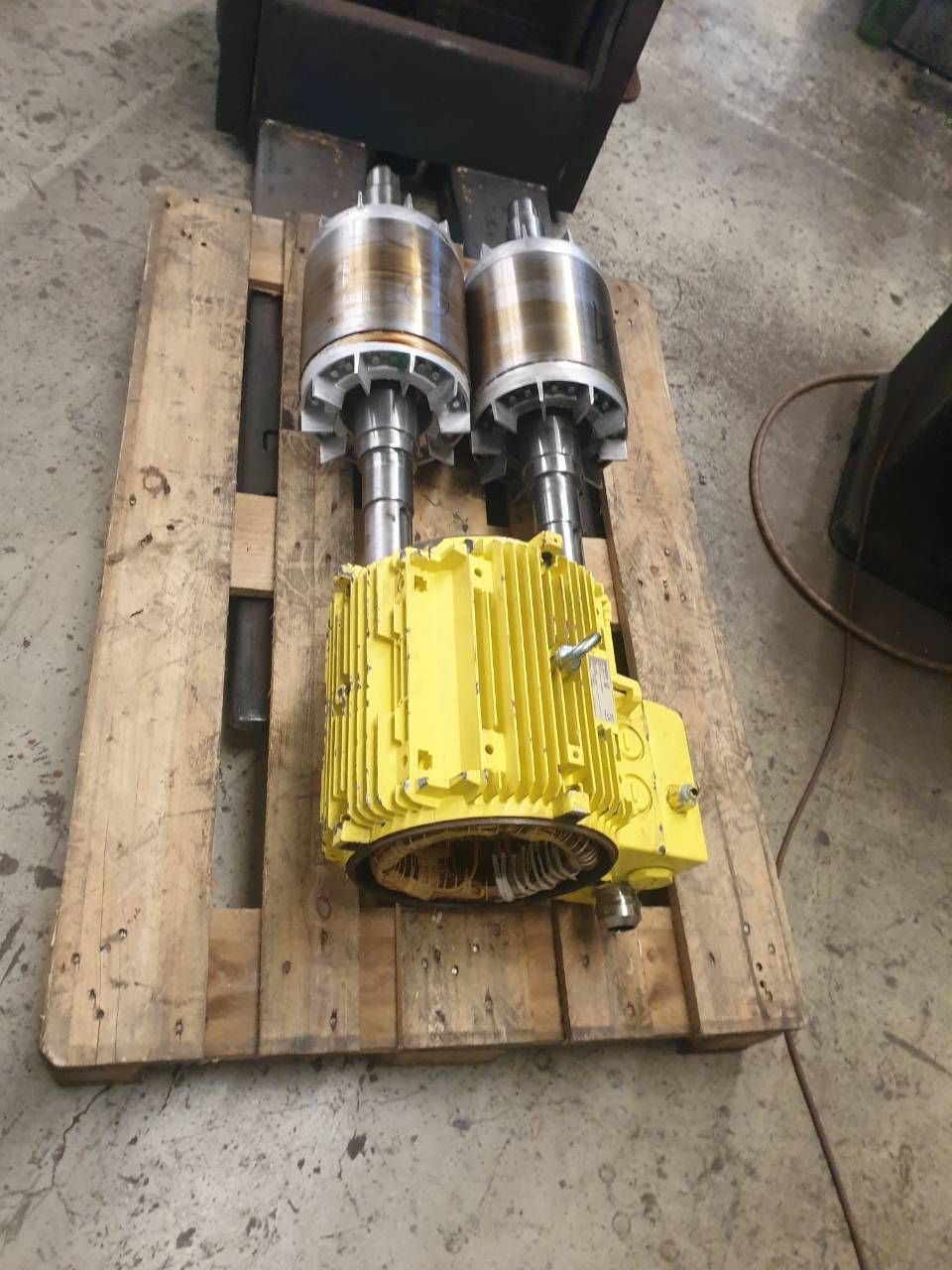 Zwei silberne Motorzylinder und ein gelbes Getriebe auf einer Holzpalette in einer Werkstatt.