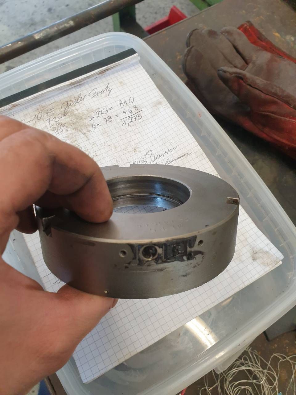 Eine Hand hält einen maschinell bearbeiteten Metallring über einer Werkbank; Papiere und ein Handschuh liegen daneben.