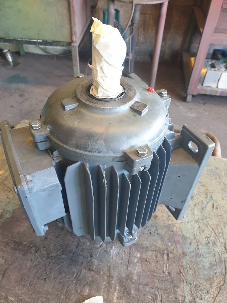 Grosser grauer Industrie-Elektromotor auf dem Werkstattboden, mit umwickeltem Kabel oben und seitlichen Montagehalterungen.