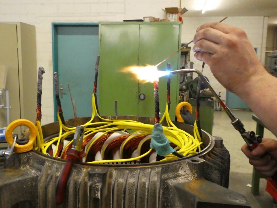 Ein Techniker lötet in einer Werkstatt Drähte an eine grosse Elektromotorspule mit einem Schweissbrenner.