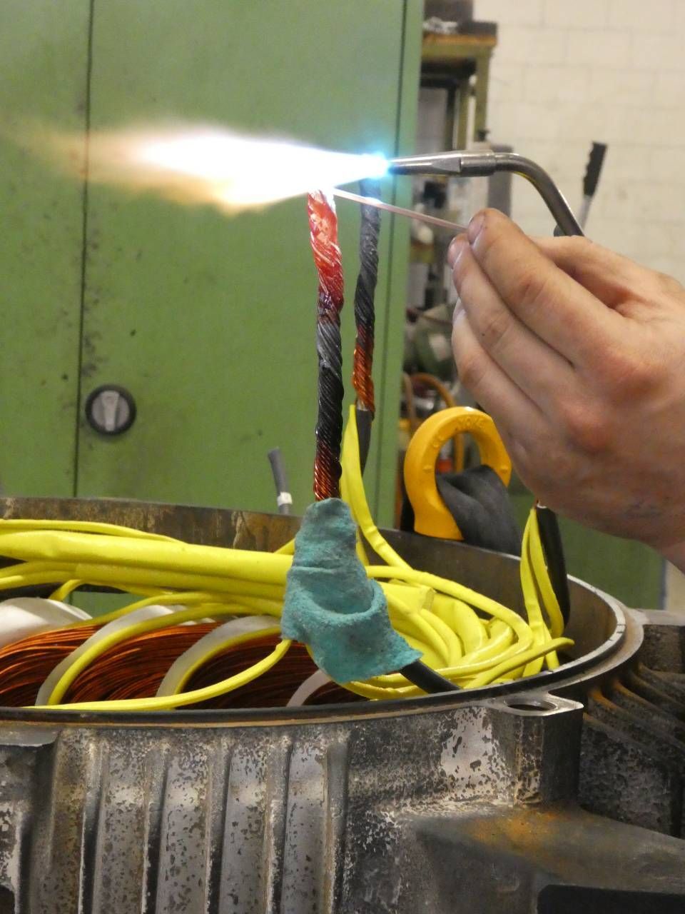 Mit einem Handbrenner werden Drähte über einem Metallbehälter mit gebündelten Kabeln und stoffumwickelten Teilen erhitzt.