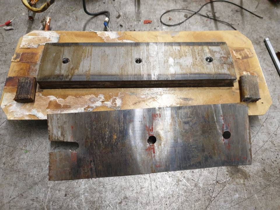 Zwei rostige Metallmaschinenteile liegen auf dem Werkstattboden: eine lange Platte und eine rechteckige Platte mit Löchern.