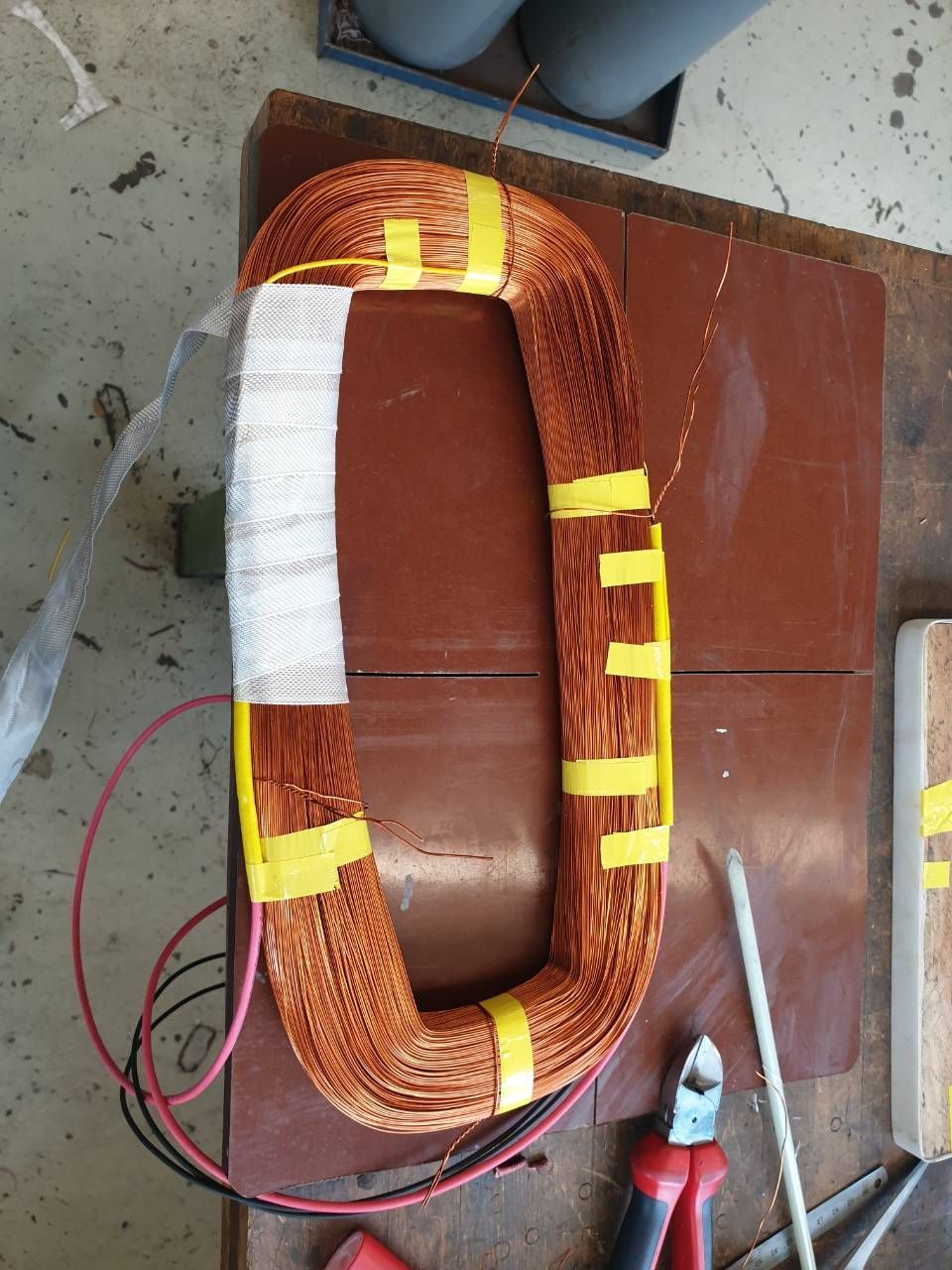Eine mit gelbem Klebeband umwickelte Kupferspule liegt auf einem braunen Tisch, daneben befinden sich rote und schwarze Drähte und rechts ein Werkzeug.